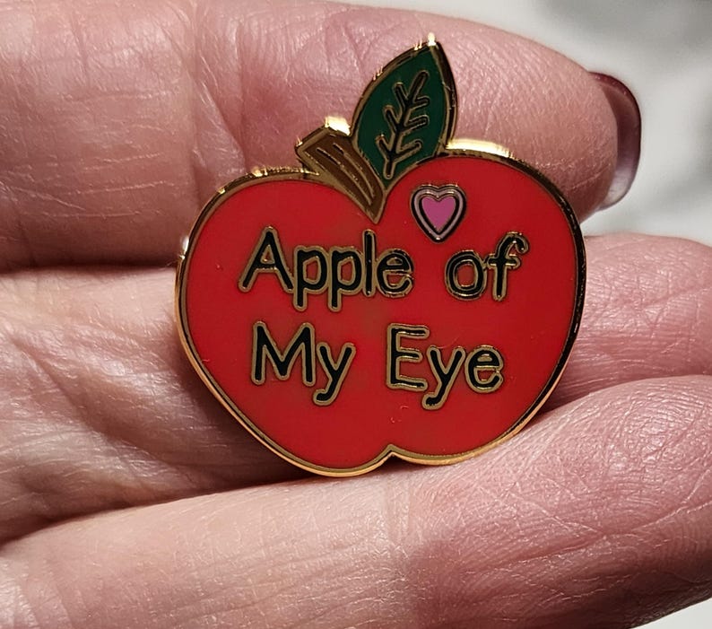 1 inch enamel pin in the shape of an apple.  Red with a small pink heart at top for Valentines.  Green leaf and Brown Stem. Shiny gold background and back. Smooth.  Hard Enamel.  Text on apple reads, "Apple of My eye."