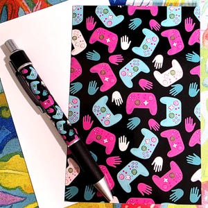 Puede incluir: Un bolígrafo negro con punta plateada y un patrón de mandos de videojuegos rosas y azules. También se ve una tarjeta de felicitación a juego con el mismo patrón de mando y contornos de manos blancas.