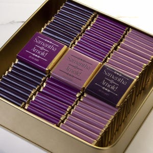 May include: A gold-colored rectangular tin filled with individually wrapped chocolate bars. The wrappers are various shades of purple, with some featuring the text "Samantha Arnold" in white. The chocolates are arranged neatly inside the tin.