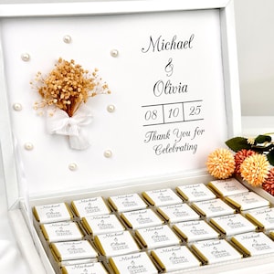 Op de afbeelding: Een witte doos gevuld met individueel verpakte chocolaatjes. De doos is versierd met parels, een klein boeket en de namen "Michael & Olivia", een datum en de zin "Thank You for Celebrating".