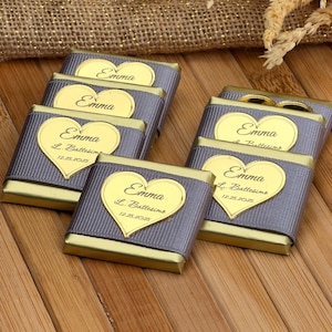Può includere: Diverse piccole barrette di cioccolato avvolte con bordi in lamina d'oro e nastro grigio. Ogni barretta ha un'etichetta dorata a forma di cuore con il nome "Emma" e la data "12.25.2025". Le barrette sono disposte su una superficie di legno.