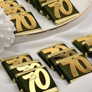 Könnte beinhalten: Nahaufnahme von Schokoriegeln mit goldenen Details. Jeder Riegel hat die goldene Zahl 70, mit dem Aufdruck "Happy Birthday Daphne" auf dem Etikett. Die Riegel sind in dunkelgrünen Stoff gewickelt und auf einem weißen Tablett angeordnet.