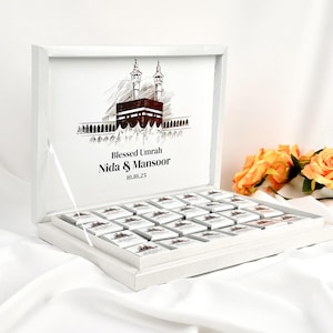 May include: A white gift box with a hinged lid, filled with small, square chocolates. The inside lid features a sketch of a religious structure with the words "Blessed Umrah Nida & Mansoor 10.10.25". The box is on a white surface.