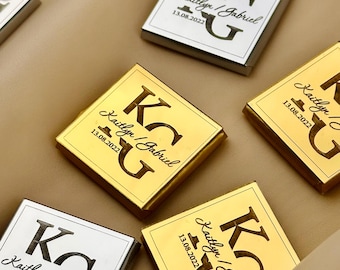 Op de afbeelding: Vierkante chocolade gunsten met goud en zilverfolie wrappers. Elke gunst is bedrukt met de initialen "KG", de namen "Kaitlyn / Gabriel" en de datum "13.08.2022". De gunsten zijn gerangschikt op een neutrale ondergrond.