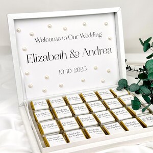 Op de afbeelding: Een witte doos gevuld met individueel verpakte chocolaatjes. Op het deksel staat "Welcome to Our Wedding", de namen "Elizabeth & Andrea" en de datum "10-10-2025". Kleine parels zijn ook zichtbaar.