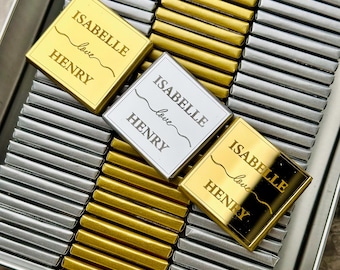 Op de afbeelding: Een zilveren en gouden blik gevuld met kleine chocolade reepjes verpakt in zilver en gouden folie. Elke reep heeft een gepersonaliseerd etiket met de tekst "Isabelle love Henry".