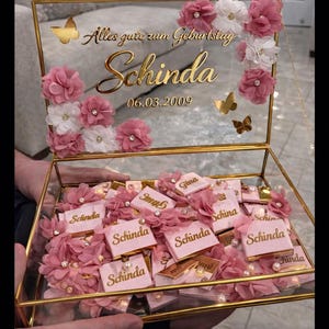 May include: A clear glass box with a gold frame, filled with pink chocolates and decorative flowers. The box is adorned with gold butterflies and the inscription "Alles gute zum Geburtstag Schinda 06.03.2009".