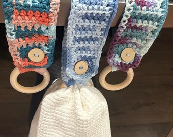 Puede incluir: Tres colgadores de toallas de cocina de ganchillo en tonos azules, coral y morado, cada uno con un anillo de madera y cierre de botón. Una toalla blanca de tejido de gofre cuelga de uno de los colgadores.