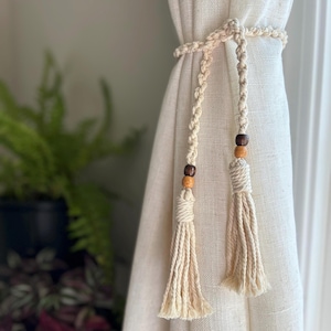 May include: Two white macrame curtain tiebacks with wooden beads. The tiebacks are tied around a white curtain.