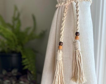 May include: Two white macrame curtain tiebacks with wooden beads. The tiebacks are tied around a white curtain.