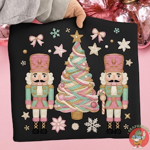 May include: Black graphic featuring a Christmas tree and two nutcrackers in pink, teal, and gold. The design includes stars, snowflakes, and bows. The nutcrackers have mustaches and hold candy canes.