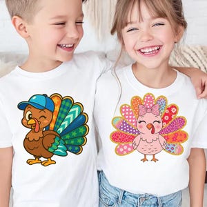 May include: Two white t-shirts with cartoon turkey designs. One shirt features a brown turkey wearing a blue cap with a colorful tail. The other shirt has a pink turkey with a bow and a patterned tail. Both shirts are for kids.