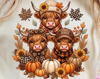 May include: A cream-coloured t-shirt featuring a graphic of three Highland cows. The cows wear a knit hat, a leopard print bow, and a baseball cap with the text FALL VIBES. They are surrounded by pumpkins, sunflowers, and autumn leaves.