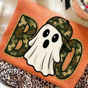 以下が含まれることがあります： オレンジ色のハロウィーンをテーマにしたスウェットシャツ。迷彩柄の「BOO」の文字が金色のグリッターで縁取られています。迷彩柄の野球帽をかぶった漫画の幽霊がデザインの中央にいます。スウェットシャツは折りたたまれています。