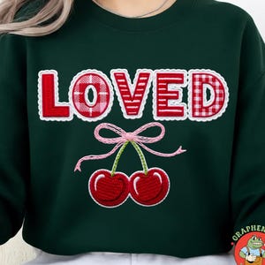 May include: Dark green sweatshirt featuring the word "LOVED" in red and white embroidered letters. The "O" has a pink plaid design. Below is an embroidered pair of red cherries with a pink bow.