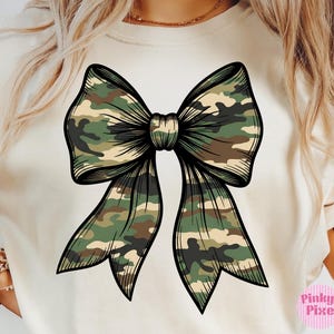 May include: A cream-colored t-shirt featuring a large, centered graphic of a bow. The bow is decorated with a green, brown, and tan camouflage pattern and outlined in bold black lines. The design includes a small pink logo in the bottom corner.