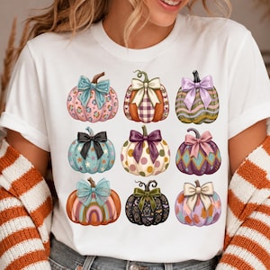 May include: A white short-sleeve t-shirt featuring a graphic print of nine pumpkins arranged in a grid. Each pumpkin displays a unique pattern, including leopard print, plaid, stripes, polka dots, and stars, and is topped with a decorative bow.