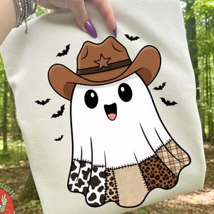 Op de afbeelding: Een crèmekleurige sweatshirt met een cartoongeest met een bruine cowboyhoed met een ster. De geest heeft een patchwork-ontwerp met koeienprint, luipaardprint en ruitpatronen. Zwarte vleermuissilhouetten omringen de geest.