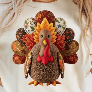 Könnte beinhalten: Cremefarbenes T-Shirt mit einem detaillierten Truthahndesign. Der Truthahn hat einen braunen Körper, einen roten Wamm und einen Fächer aus bunten Federn in Braun-, Orange- und Gelbtönen. Das Design ist auf dem Shirt zentriert und schafft ein festliches Herbstthema.