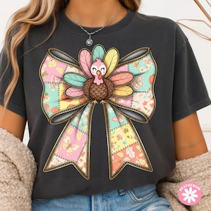 May include: Dark gray t-shirt featuring a colorful turkey design. The turkey is centered on a large bow with a patchwork pattern in pastel colors. The bow has a gold outline, and the turkey has a pink head and colorful feathers.