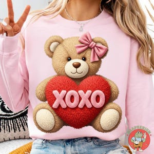 May include: Pink sweatshirt featuring a teddy bear holding a red heart with "XOXO" in white letters. The teddy bear has a pink bow and is light brown. The sweatshirt is a solid pink color.