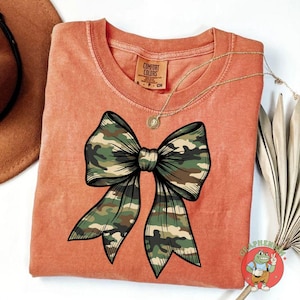 Puede incluir: Camiseta naranja Comfort Colors con un gran dise&ntilde;o de lazo de camuflaje con contorno negro. Un collar dorado con un colgante est&aacute; drapeado sobre la camiseta. La camiseta tiene una etiqueta que dice "Comfort Colors".