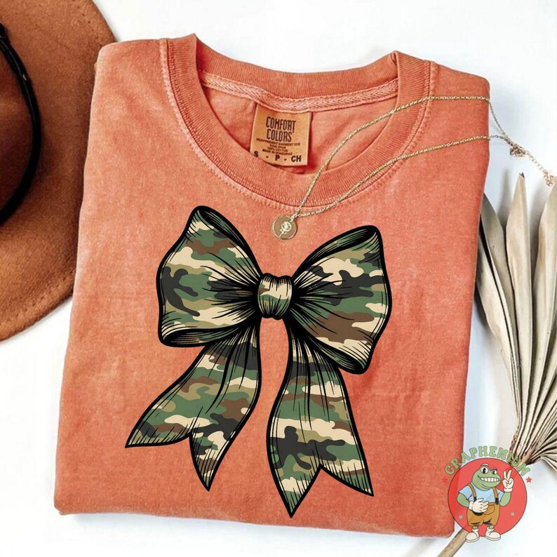 Puede incluir: Camiseta naranja Comfort Colors con un gran dise&ntilde;o de lazo de camuflaje con contorno negro. Un collar dorado con un colgante est&aacute; drapeado sobre la camiseta. La camiseta tiene una etiqueta que dice "Comfort Colors".