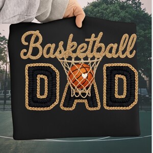 May include: Black pillow with the words "Basketball DAD" in a woven design. The word "Basketball" is in a light brown color, and the word "DAD" is in black with a light brown border. A basketball is in the net.