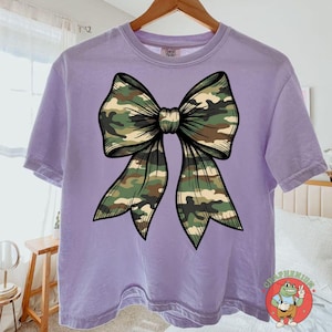 Puede incluir: Camiseta lila con un gran dise&ntilde;o de lazo de camuflaje. El lazo est&aacute; centrado y tiene un contorno detallado. La camiseta de manga corta tiene un corte relajado, ideal para un look casual.