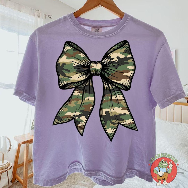 Puede incluir: Camiseta lila con un gran dise&ntilde;o de lazo de camuflaje. El lazo est&aacute; centrado y tiene un contorno detallado. La camiseta de manga corta tiene un corte relajado, ideal para un look casual.