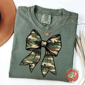 Puede incluir: Camiseta verde salvia con un gran dise&ntilde;o de lazo de camuflaje. El lazo est&aacute; delineado en negro. Un collar plateado con un colgante est&aacute; drapeado sobre la camisa. La camisa tiene una etiqueta Comfort Colors.