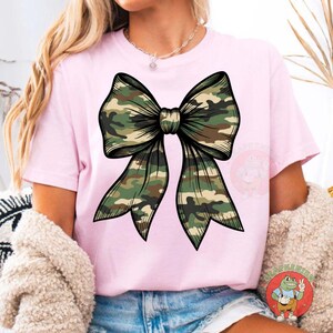 Puede incluir: Camiseta rosa con un gran dise&ntilde;o de lazo en patr&oacute;n de camuflaje. El lazo, con contorno detallado, est&aacute; centrado. El estampado de camuflaje incluye tonos verdes, marrones y beige.