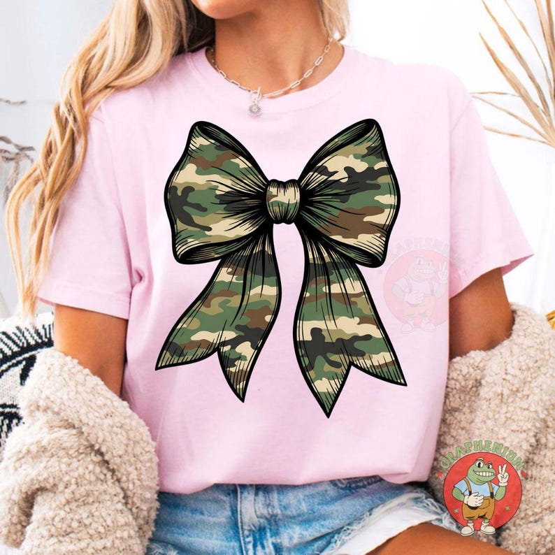 Puede incluir: Camiseta rosa con un gran dise&ntilde;o de lazo en patr&oacute;n de camuflaje. El lazo, con contorno detallado, est&aacute; centrado. El estampado de camuflaje incluye tonos verdes, marrones y beige.
