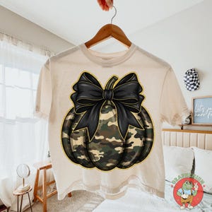 May include: A cream-colored t-shirt featuring a pumpkin design with a black bow and camouflage pattern. The pumpkin has a gold outline. The shirt is hanging on a wooden hanger.