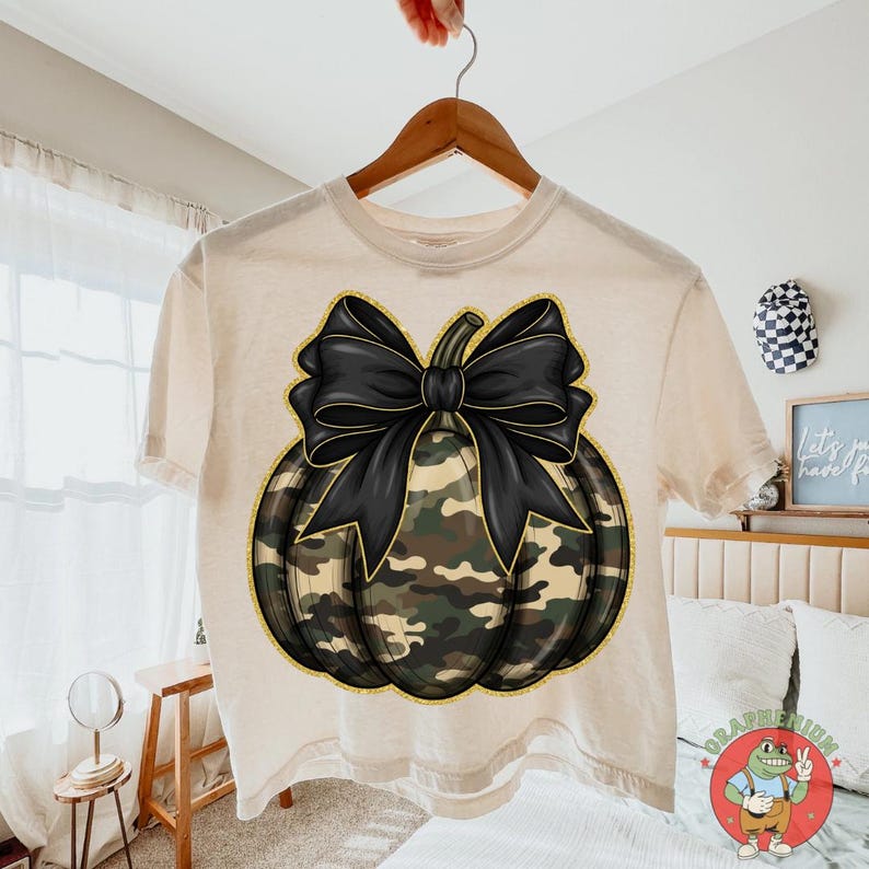 May include: A cream-colored t-shirt featuring a pumpkin design with a black bow and camouflage pattern. The pumpkin has a gold outline. The shirt is hanging on a wooden hanger.