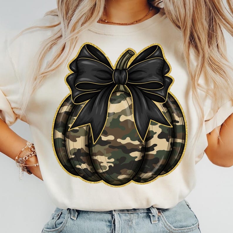 May include: Cream-colored t-shirt featuring a camouflage pumpkin design with a black bow and gold glitter trim. The pumpkin has a stem and is the focal point of the shirt. The design is centered on the front of the shirt.
