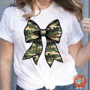 Puede incluir: Camiseta blanca con un dise&ntilde;o de lazo grande en camuflaje. El lazo presenta contornos detallados y una textura realista, con tonos verdes, marrones y beige. La camiseta es de cuello redondo cl&aacute;sico.