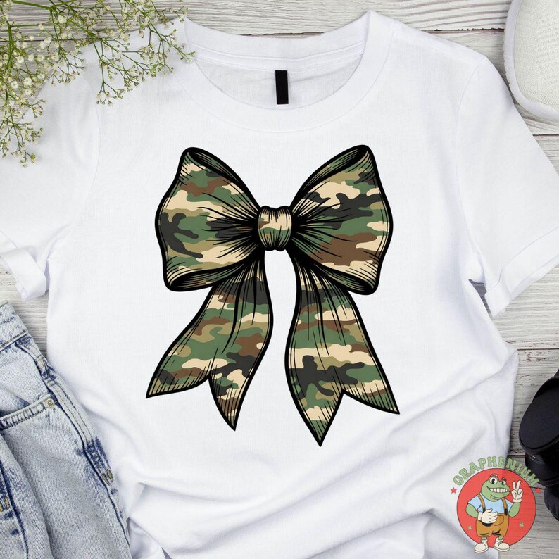 Puede incluir: Camiseta blanca con un gran lazo estampado. El lazo presenta un patr&oacute;n de camuflaje en tonos verdes, marrones y beige, delineado en negro. El dise&ntilde;o est&aacute; centrado en la camiseta, creando un impacto visual audaz.