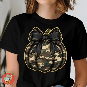 May include: Black t-shirt featuring a camouflage pumpkin design with a large black bow. The pumpkin has a gold outline. The design is centered on the shirt, creating a festive and stylish look.