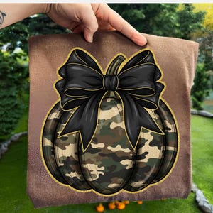 May include: A decorative square pillow featuring a camouflage pumpkin design with a large black bow. The pumpkin has a gold glitter outline and stem. The pillow is held up by a hand.