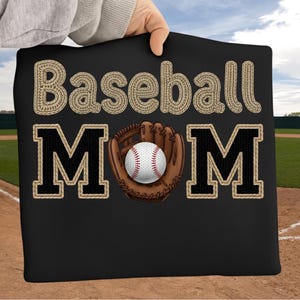 May include: Black pillow with the words "Baseball Mom" in tan and black lettering. A baseball glove with a baseball in the center is also featured. The pillow is being held up by a person.