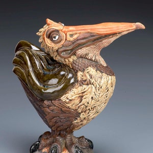 May include: A ceramic sculpture of a pelican with a brown, green, and orange glaze. The pelican has a long beak and large eyes. The sculpture is standing on a base with a textured surface.