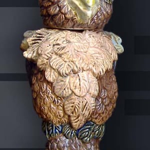 Può includere: Una statuetta di uccello in ceramica con dettagli marroni, dorati e verdi. L'uccello ha un becco dettagliato e grandi occhi rotondi. La statuetta è in piedi su una base con tre artigli.