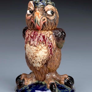 Può includere: Una statuetta di gufo in ceramica con un cappello blu e verde, piume marroni e un petto con motivo rosso e marrone. Il gufo ha grandi occhi rotondi e un lungo becco appuntito. Il gufo è in piedi su una base blu e verde con artigli neri.