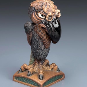 May include: A detailed ceramic bird figurine with a stern expression. The bird has a black beak, brown and black feathers, and is perched on a square base with green trim. The figurine is approximately 15 cm tall.
