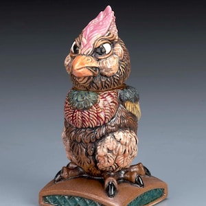 Può includere: Una stravagante statuina di uccello in ceramica con cresta rosa, becco arancione e piume dettagliate nei toni del marrone, rosso e verde. L'uccello è posato su un cuscino marrone con un bordo verde a motivi. Un oggetto decorativo unico.