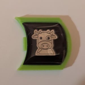 May include: A green plastic key chain with a black and silver cow design. The cow has a white spot on its face and is outlined in silver.