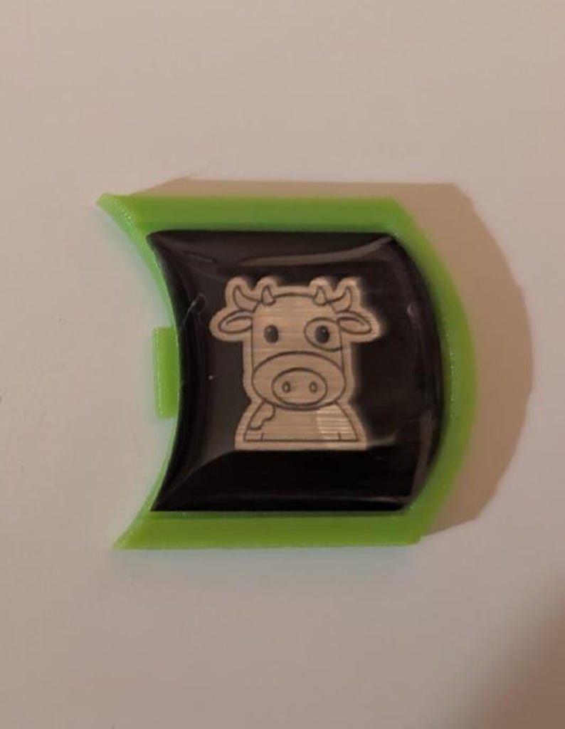 May include: A green plastic key chain with a black and silver cow design. The cow has a white spot on its face and is outlined in silver.