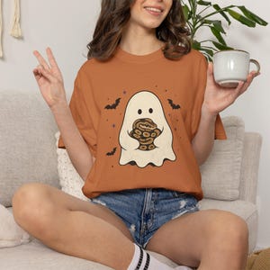 May include: A burnt orange short-sleeve t-shirt featuring a graphic print of a white ghost holding a brown patterned snake. The design includes small black bats and purple stars surrounding the ghost. The shirt has a crew neckline and a relaxed fit.