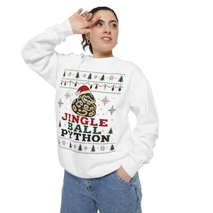 May include: A white long-sleeve sweatshirt featuring a graphic of a ball python wearing a Santa hat, surrounded by a holiday-themed pattern of Christmas trees, snowflakes, and string lights, with the text JINGLE BALL PYTHON printed in red, green, and black.